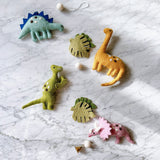 The Jurassic Dinosaur felt garland is displayed vertically on a marble surface. This playful felt garland features a light blue Stegosaurus with dark blue spikes, a yellow Brontosaurus, a green Tyrannosaurus Rex, and a pink Triceratops, each adorned with white and brown dots. Two green monstera leaves and white felt balls separate the elements. A small copper bell dangles charmingly at the garland's end.