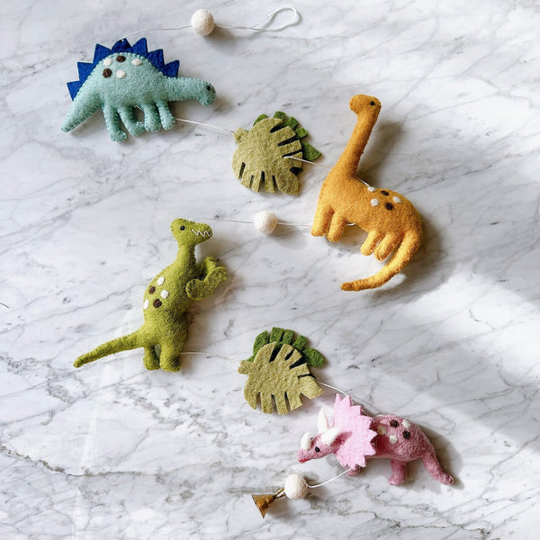 The Jurassic Dinosaur felt garland is displayed vertically on a marble surface. This playful felt garland features a light blue Stegosaurus with dark blue spikes, a yellow Brontosaurus, a green Tyrannosaurus Rex, and a pink Triceratops, each adorned with white and brown dots. Two green monstera leaves and white felt balls separate the elements. A small copper bell dangles charmingly at the garland's end.
