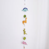 A Jurassic dinosaur felt garland hangs vertically against a plain white wall, creating a playful atmosphere. The felt garland features a variety of felt dinosaurs and monstera leaves, separated by small white felt balls. From top to bottom, it showcases a light blue Stegosaurus, a green monstera leaf, an orange Brontosaurus, a light green T-Rex, another green monstera leaf, and a pink Triceratops. A small copper bell is attached at the very bottom.
