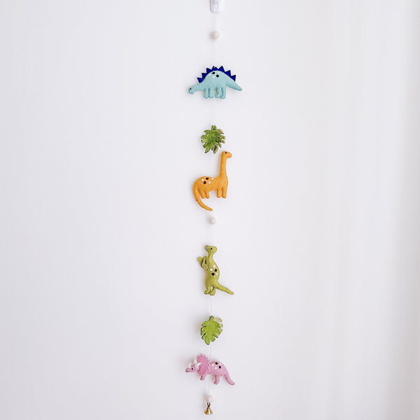 A Jurassic dinosaur felt garland hangs vertically against a plain white wall, creating a playful atmosphere. The felt garland features a variety of felt dinosaurs and monstera leaves, separated by small white felt balls. From top to bottom, it showcases a light blue Stegosaurus, a green monstera leaf, an orange Brontosaurus, a light green T-Rex, another green monstera leaf, and a pink Triceratops. A small copper bell is attached at the very bottom.