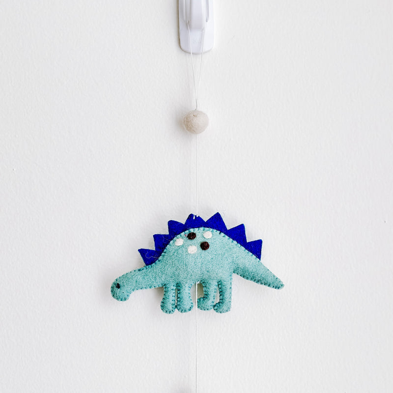A light blue felt Jurassic Dinosaur garland, featuring a Stegosaurus, hangs vertically on a white wall, affixed by a white adhesive hook. The detailed felt Stegosaurus has dark blue spikes and small stitches around its edges. A round felt ball is positioned above the Stegosaurus, and a charming copper bell hangs at the garland's end.