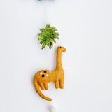 A vertical felt Jurassic dinosaur garland is displayed against a clean white background. It features a grey felt brontosaurus with a long neck and tail, adorned with white and black spots. Above it is a vibrant green monstera leaf, connected by a thin white string. A fluffy white bell hangs at the bottom, adding a charming, playful touch.