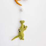 A section of a Jurassic dinosaur felt garland is displayed vertically against a bright white wall. It features a partially visible green felt monstera leaf and an off-white felt ball at the top. Below, a vibrant green felt T-Rex dinosaur hangs, detailed with small white spots, a stitched mouth, and raised arms. The felt garland is suspended by a thin white string, creating a playful and educational display.
