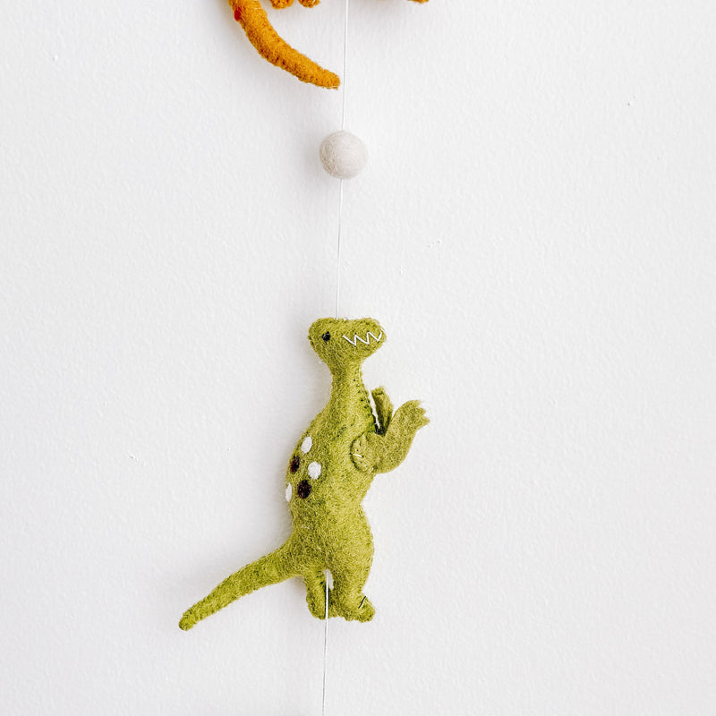 A section of a Jurassic dinosaur felt garland is displayed vertically against a bright white wall. It features a partially visible green felt monstera leaf and an off-white felt ball at the top. Below, a vibrant green felt T-Rex dinosaur hangs, detailed with small white spots, a stitched mouth, and raised arms. The felt garland is suspended by a thin white string, creating a playful and educational display.
