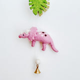 A close-up of a Jurassic Dinosaur felt garland hangs vertically against a white wall. This section features a vibrant green monstera leaf, a pink triceratops with white horns and brown spots, followed by a beige felt ball, and a copper bell at the bottom, creating a playful and whimsical design.