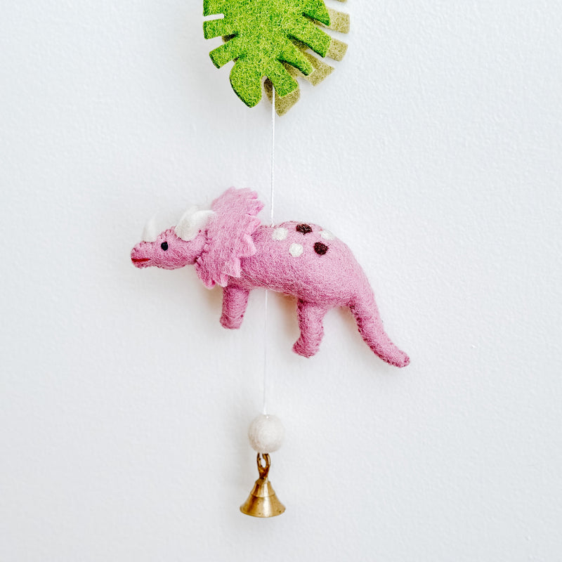 A close-up of a Jurassic Dinosaur felt garland hangs vertically against a white wall. This section features a vibrant green monstera leaf, a pink triceratops with white horns and brown spots, followed by a beige felt ball, and a copper bell at the bottom, creating a playful and whimsical design.