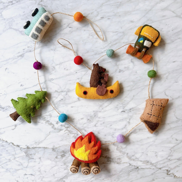 A playful outdoor adventure felt garland is arranged in a semi-circular display on a white marble background. The felt garland features charming figures on a brown string, interspersed with colorful felt balls. Figures include a light blue mini van, green tree, red and yellow campfire, tan tent, yellow kayak with a brown bear, and a yellow and brown backpack. Felt balls are in orange, red, purple, blue, and green.