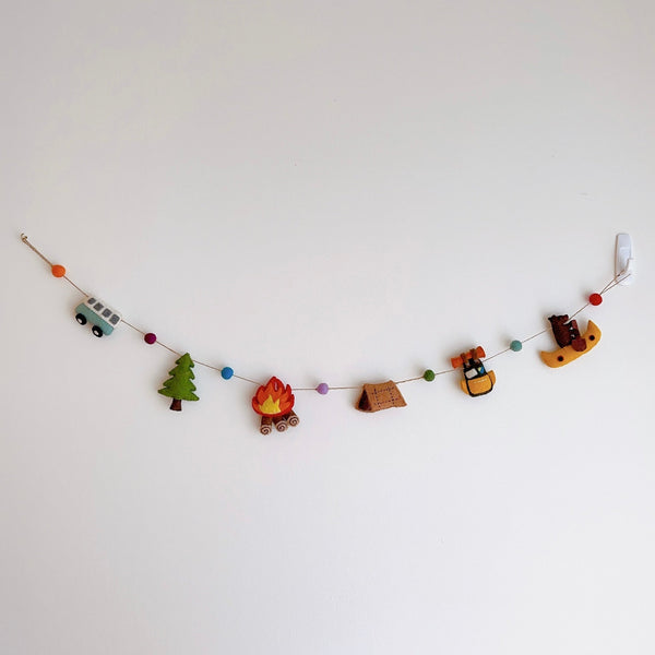 An Outdoor Adventure felt garland hangs against a plain white wall, held by a white hook. This playful felt garland features colorful camping-themed elements crafted from felt, including a blue mini van, a green tree, a campfire with orange flames, a brown tent, a yellow backpack, and a yellow kayak with a brown bear. The elements are strung along a thin brown string with colorful felt balls spaced in between.