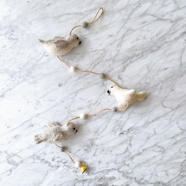 A felt seal garland is displayed on a white marble surface, exuding a calm and playful atmosphere. The garland features three soft felt seals—two light grey and one white—interspersed with alternating white and grey felt balls on a brown string. The bottom felt seal is adorned with a small copper bell.