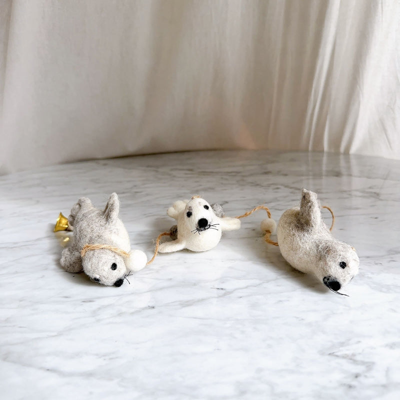 A felt seal garland, featuring three charming wool felt seals, rests on a white marble surface against an off-white fabric backdrop. The seals are connected by rustic twine interspersed with fuzzy white balls. The light grey seal on the left has a copper bell and white pom-pom, the center seal is creamy white, and the light grey one on the right faces downward. Each seal has black eyes and whiskers, creating a cute marine-themed decoration.