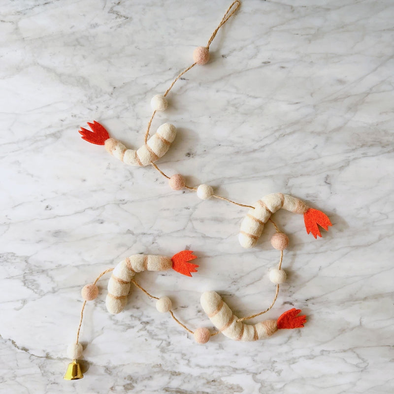 A playful Felt Shrimp Garland rests on a clean white marble surface. It features three white felt shrimp with light tan stripes and bright orange tails, evenly spaced along a thin twine. Small, round felt balls in light pink and white are interspersed between the shrimp, and a single copper bell adorns one end.