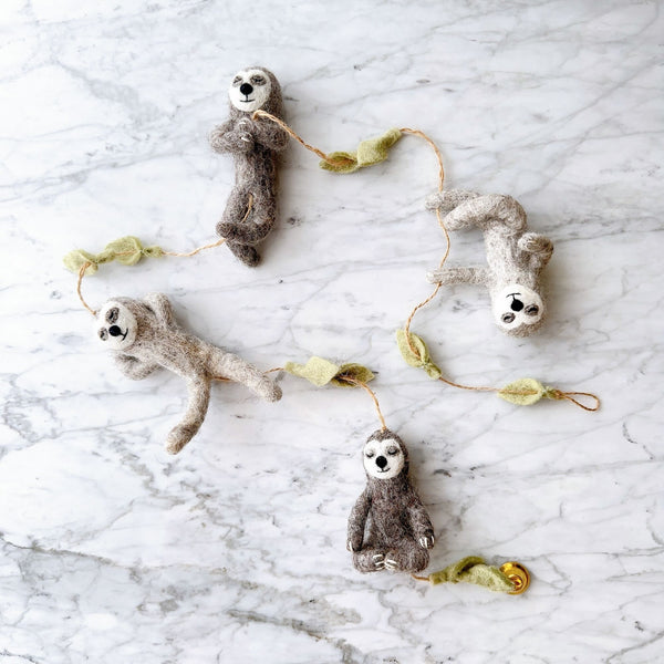 A whimsical felt garland, featuring four grey and brown sloths and light green felt leaves, rests on a white marble surface. This serene sloth felt garland showcases sloths in various playful poses, including hanging upside down, meditating, reaching out, and standing upright, all connected by thin twine.