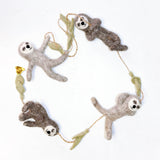 A playful wool felt sloth garland is displayed against a white background. The garland features four poseable sloths, crafted from light and dark brown felt with white faces and black embroidered features. They are strung on a brown twine with light green felt branch segments separating each sloth.