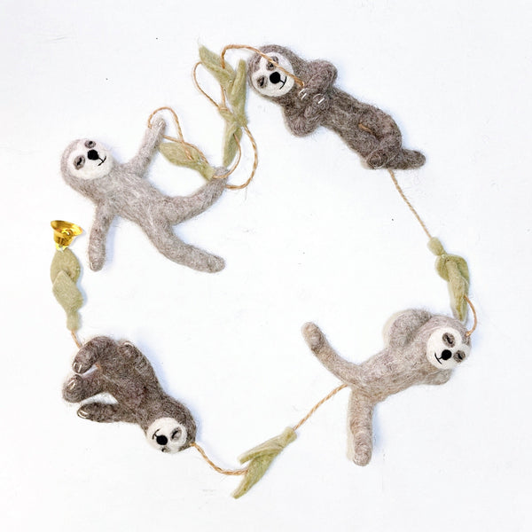 A playful wool felt sloth garland is displayed against a white background. The garland features four poseable sloths, crafted from light and dark brown felt with white faces and black embroidered features. They are strung on a brown twine with light green felt branch segments separating each sloth.
