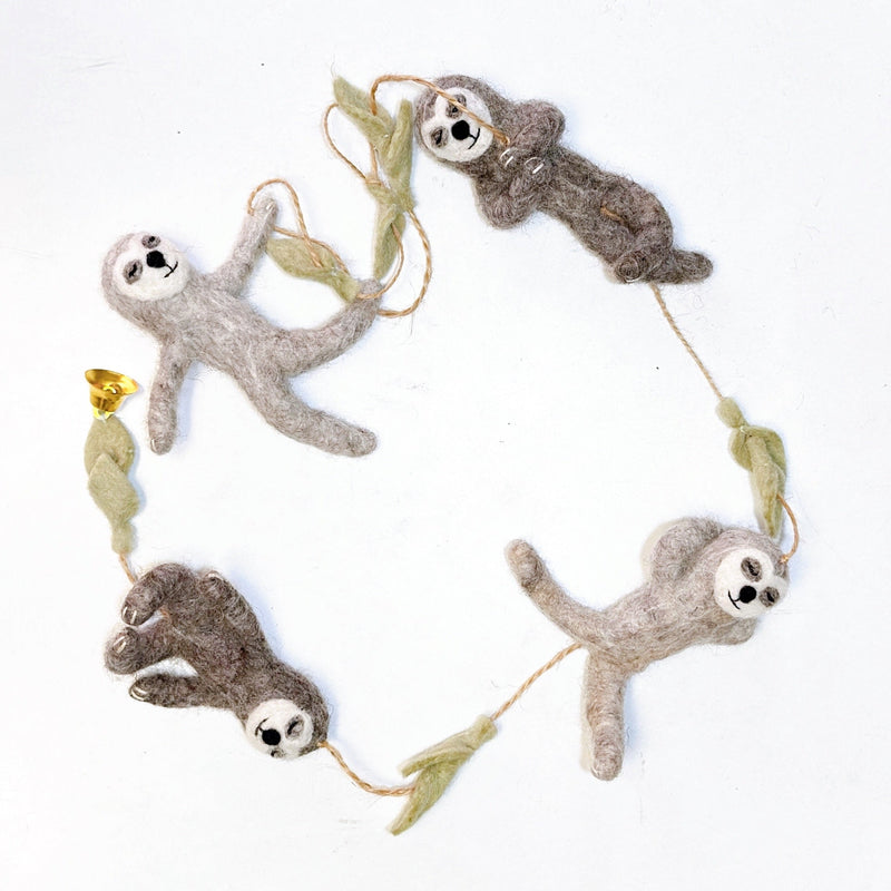 A playful wool felt sloth garland is displayed against a white background. The garland features four poseable sloths, crafted from light and dark brown felt with white faces and black embroidered features. They are strung on a brown twine with light green felt branch segments separating each sloth.