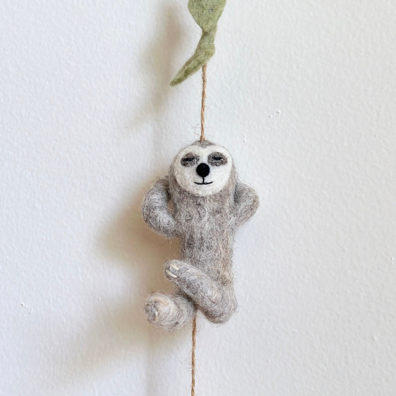 A gray wool felt sloth, part of the Slothful Serenity felt garland, hangs suspended by a natural twine against a white wall. The soft wool felt sloth features a peaceful expression with closed eyes and a gentle smile, its arms raised in a relaxed pose. A small green felt leaf is attached to the twine above the sloth.
