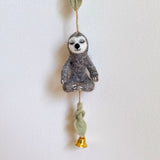 A detailed view of the Felt Slothful Serenity garland is presented against a bright white wall, creating a calm and cheerful scene. The gray felt sloth is in a seated yoga pose, suspended from a thin brown string. It has a white face with closed eyes, in a meditating position, with its posable limbs shaped by hidden wires.