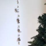 A calming felt sloth garland is suspended against a clean white wall, with a green Christmas tree partially visible on the right. This unique handmade felt garland features four gray sloths with posable limbs, separated by light green felt leaves, strung together on a brown string. A small copper bell adds a charming accent at the garland's end.