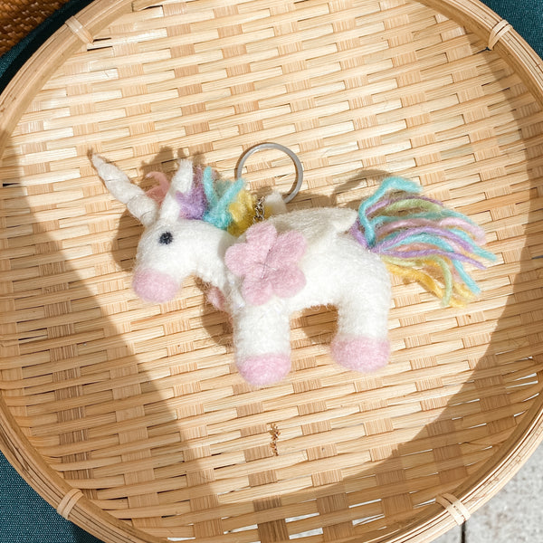 A pastel unicorn felt keychain is displayed on a round, woven straw tray, set against a corner of a teal surface. The white felt unicorn features a pink snout and hooves, a pastel yellow, blue, and purple yarn mane and tail, and a pink felt flower embellishment. A metal keychain with clasp is attached to the top. The woven texture of the tray adds a rustic touch to this charming handmade felt keychain.