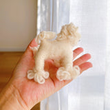 A handmade white poodle felt keychain is gently presented by a female hand against a light, airy background featuring sheer white curtains. This charming wool felt keychain, in a soft off-white hue, features a rounded body, fluffy legs, and a curly head, giving it a soft, textured appearance.