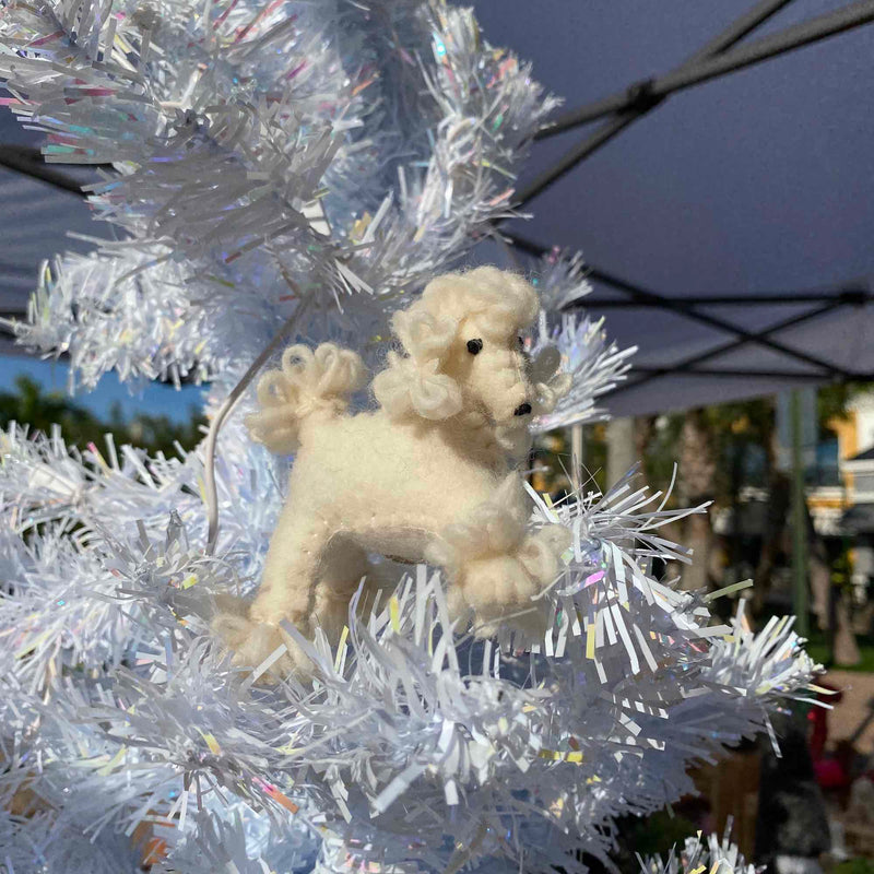 A white poodle felt keychain is nestled among the branches of an artificial white Christmas tree adorned with shiny tinsel. The off-white needle felted poodle features a curly wool texture, detailed with small black eyes and a nose.