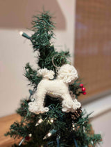 A white poodle felt keychain hangs on a small, decorated Christmas tree adorned with tiny white lights and red berries. This handmade felt keychain features a textured, fluffy white body with rounded shapes, crafted from 100% wool felt. A metal key ring is visible at the top, attaching it to a tree branch. The background shows a blurred neutral wall and wood, creating a warm, festive atmosphere.