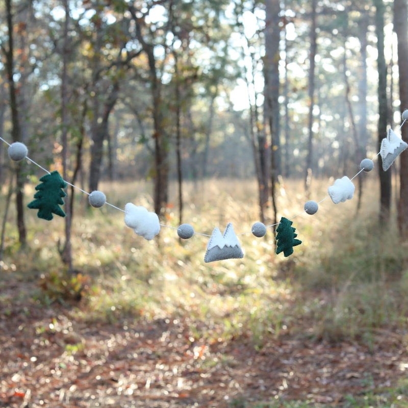 The wool felt mountain and cloud garland is suspended, set against a sunlit forest backdrop, evoking a sense of nature. This 3D-designed garland features alternating green felt trees, gray and white felt mountains, and soft felt clouds. Each element is separated by small gray felt balls, creating a charming display.