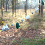 A wool felt garland, featuring mountains and clouds, is displayed against an outdoor backdrop with trees and ground covered in leaves. This garland showcases alternating 3D-designed gray and white mountains, white clouds, and green trees, separated by gray felt balls. The soft, textured wool shapes are strung on a thin white string, creating a natural, forest-like theme.