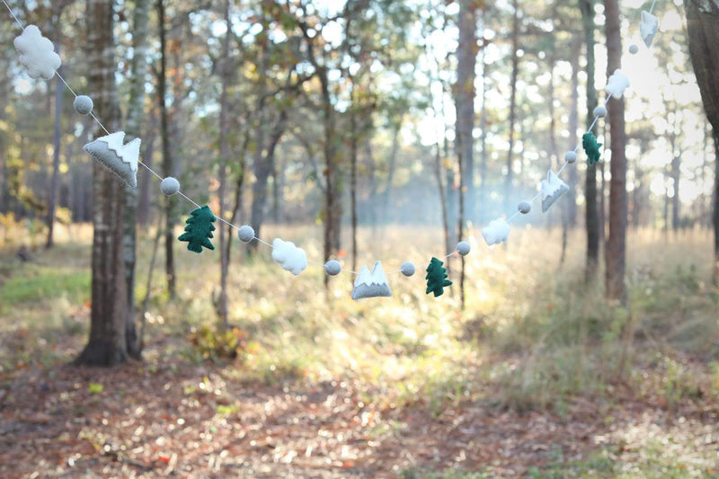 A wool felt mountain and cloud garland is strung across a blurred forest background, evoking a calm and playful atmosphere. It features alternating white clouds, grey mountains with white snow caps, and dark green fir trees. Small felt balls separate these delightful elements along the white string.