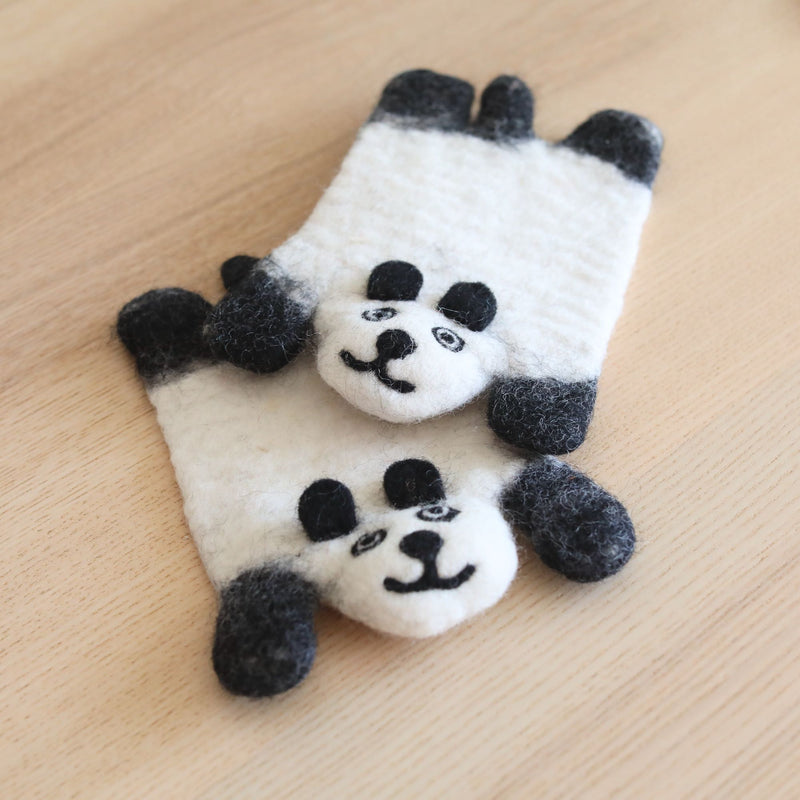 Two white wool felt panda coasters are playfully stacked on a light wooden surface, setting a cozy scene. These handcrafted panda coasters feature black felt details for ears, eyes, and limbs. Each unique felt panda coaster has a simple, smiling needle-felted face, highlighting its ethically handmade charm.