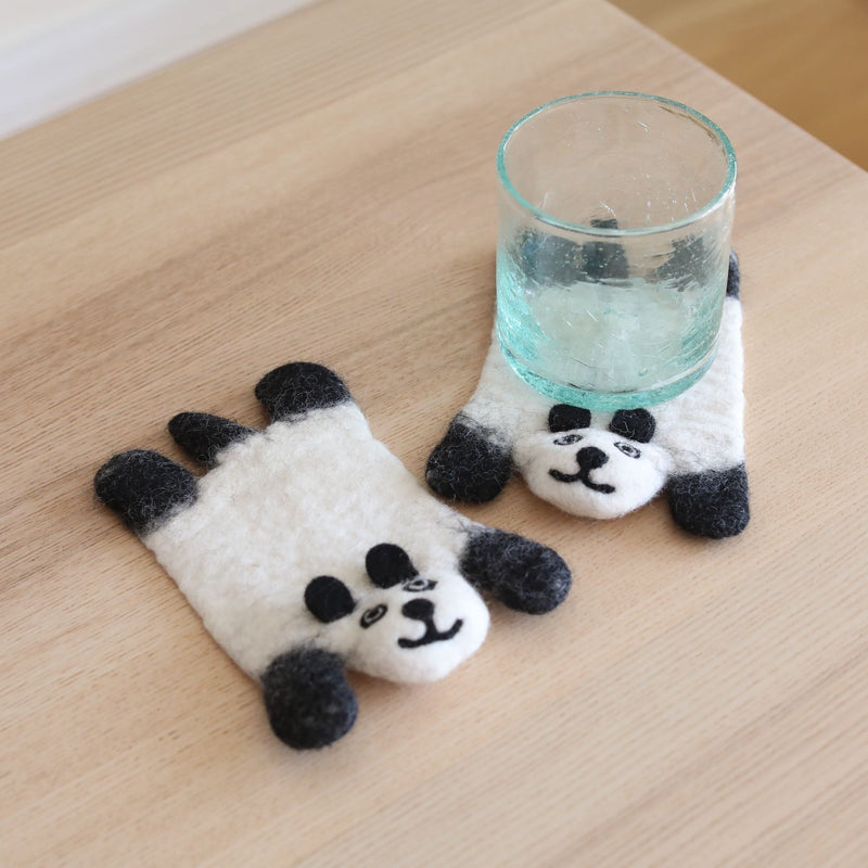 Two felt panda coasters are displayed on a light wooden surface, creating a calm and inviting atmosphere. One round felt panda coaster holds a glass of water, while the other lies flat. Each meticulously crafted coaster, made from 100% sheep wool, features a white face with black ears, muzzle, and needle-felted eyes. These unique, ethically handmade coasters support Nepalese female artisans.