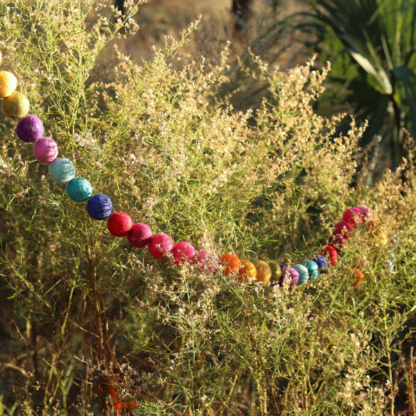 A vibrant felt rainbow pompom ball garland is draped across a leafy green bush in an outdoor setting. The colorful, spherical felt balls, in a rainbow assortment of yellow, olive green, purple, pink, teal, light blue, dark blue, and red, have a soft, fuzzy texture. Tiny white flowers dot the bush, with a palm tree in the background, creating a playful, summery atmosphere.