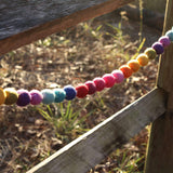 A cheerful Felt Rainbow Pompom Ball Garland is playfully draped on a rustic wooden fence in a natural outdoor setting. The garland features soft, fuzzy 100% wool felt balls, needle felted and 3D designed, in a vibrant rainbow of yellow, green, purple, pink, red, orange, and blue. This handcrafted garland adds a bright, cheerful touch.