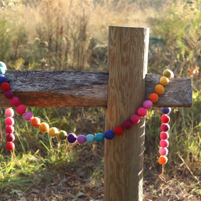 A Rainbow Pompom Ball Wool Felt Garland is gracefully draped over a weathered wooden post outdoors, set against a soft background of tall grass and foliage. The garland features a string of colorful, 3D-designed wool felt balls in vibrant rainbow hues: red, orange, yellow, green, blue, and purple. These soft, slightly fuzzy felt balls are evenly spaced, adding a playful and cheerful touch to the rustic scene.