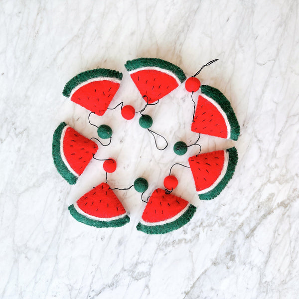 A cheerful wool felt watermelon garland is arranged in a partial circle on a white marble surface. The garland features several 3D wool felt watermelon slices with red centers, black seed stitches, thin white rinds, and green outer layers. Small red and green wool felt balls are interspersed along a black string, which connects all the elements. The bright setting is enhanced by subtle gray veining on the marble.