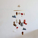 A felt woodland animal baby mobile hangs gracefully against a stark white wall, creating a calm and natural display. Handcrafted from wool felt, it features detailed 3D figures of a brown squirrel, white bunny, brown owl, gray elephant, and red fox. White clouds, blue mountains, red-capped mushrooms, and brown acorns are also suspended at varying heights, adding charm and visual balance.