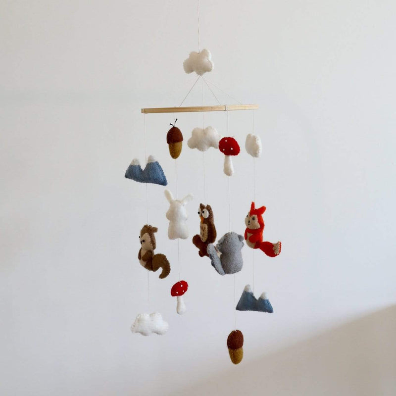 A felt woodland animal baby mobile hangs gracefully against a stark white wall, creating a calm and natural display. Handcrafted from wool felt, it features detailed 3D figures of a brown squirrel, white bunny, brown owl, gray elephant, and red fox. White clouds, blue mountains, red-capped mushrooms, and brown acorns are also suspended at varying heights, adding charm and visual balance.