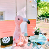 A needle felted flamingo finger puppet is held by a female hand against an outdoor backdrop featuring a retro camper. This handmade flamingo finger puppet is vibrantly pink with a white and black beak, black eyes, and a fluffy texture, ready for felt puppet finger play.