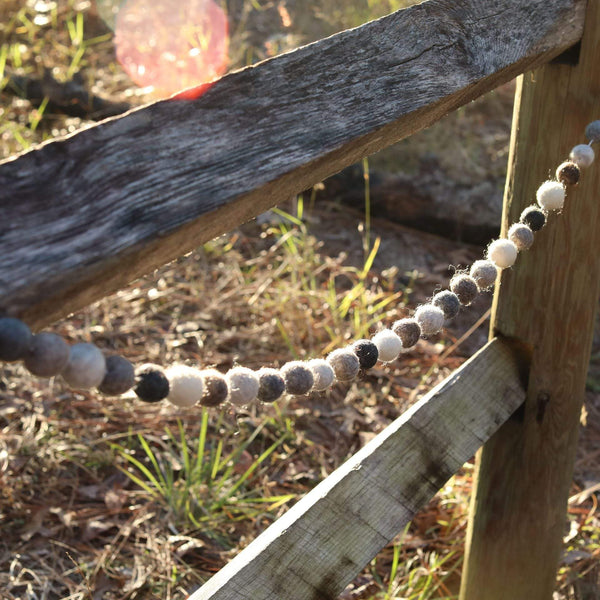 A gray wool felt ball garland is draped across a weathered wooden fence outdoors, creating a rustic and natural scene. The garland displays spherical felt balls in a gradient of light to dark gray hues. Soft sunlight filters through, highlighting the aged wood grain of the fence and the natural foliage in the background, adding warmth to the tranquil outdoor setting.