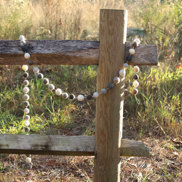 A Gray Felt Ball Garland is draped over a weathered wooden fence, set in a field of tall grass, creating a rustic outdoor scene. This felt ball garland features wool felted balls in varying shades of gray, cream, and brown, evenly spaced on a thin white string. The visible texture of the wool felt adds a soft, handcrafted feel.