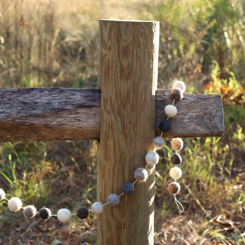 A gray wool felt ball garland is casually draped over a weathered wooden fence post, set against a sunlit, grassy field. This rustic garland features evenly spaced, spherical wool felt balls in varying shades of gray, brown, and white, adding a warm touch to the scene.