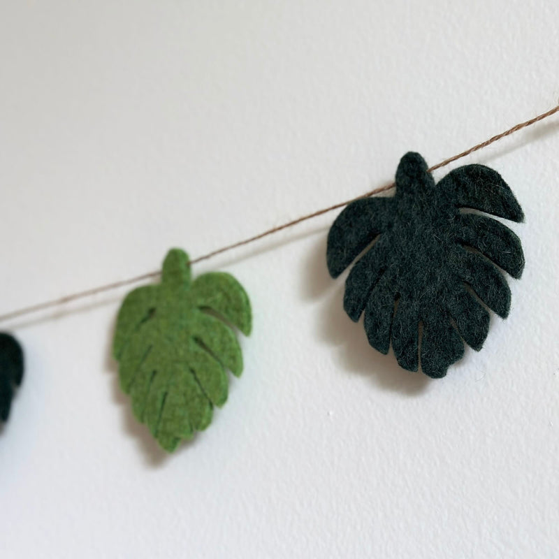 A Greenish Monstera Leaf felt garland, a nature-inspired decor piece, is shown in a close-up, hanging against a stark white wall. The felt garland features evenly spaced monstera leaves on a thin jute string. Each soft leaf alternates between light and dark green, with a textured surface mimicking natural veins.