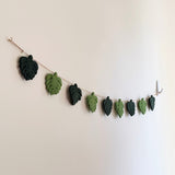 A greenish Monstera Leaf wool felt garland, a nature-inspired decor piece, is elegantly suspended against a plain, light beige wall. The garland features alternating monstera leaves in two tones of green, connected by a thin jute thread. Transparent hooks secure the garland on both sides.