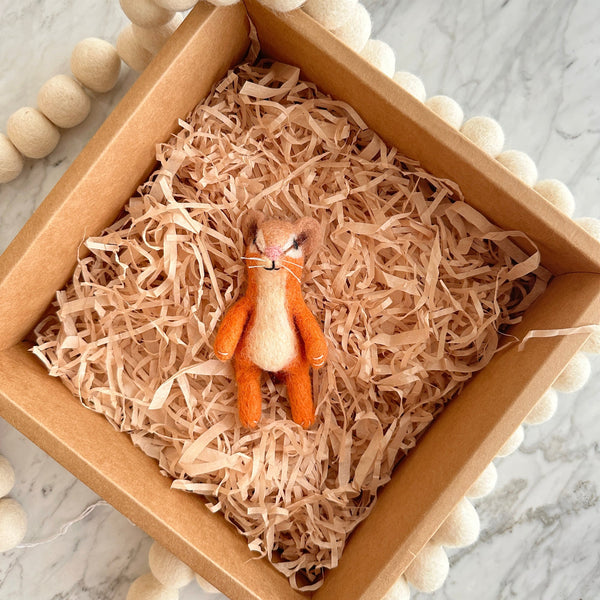 A handcrafted wool felt hamster finger puppet is presented in a square brown cardboard box filled with shredded paper, resting on a marble-like surface. This adorable hamster finger puppet features soft orange and white wool felt with tiny ears and whiskers. A white spherical wool garland adds a decorative touch within the box, creating a playful and inviting display perfect for imaginative play.