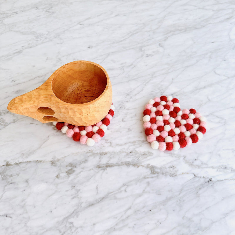 A set of four heart-shaped wool felt Love coasters, crafted from red, white, and pink organic lamb wool felt balls, rests on a bright marble surface. One handcrafted felt coaster supports a light wooden mug, while another displays its charming design. These unique, ethically made coasters reflect warmth and artisanal quality.