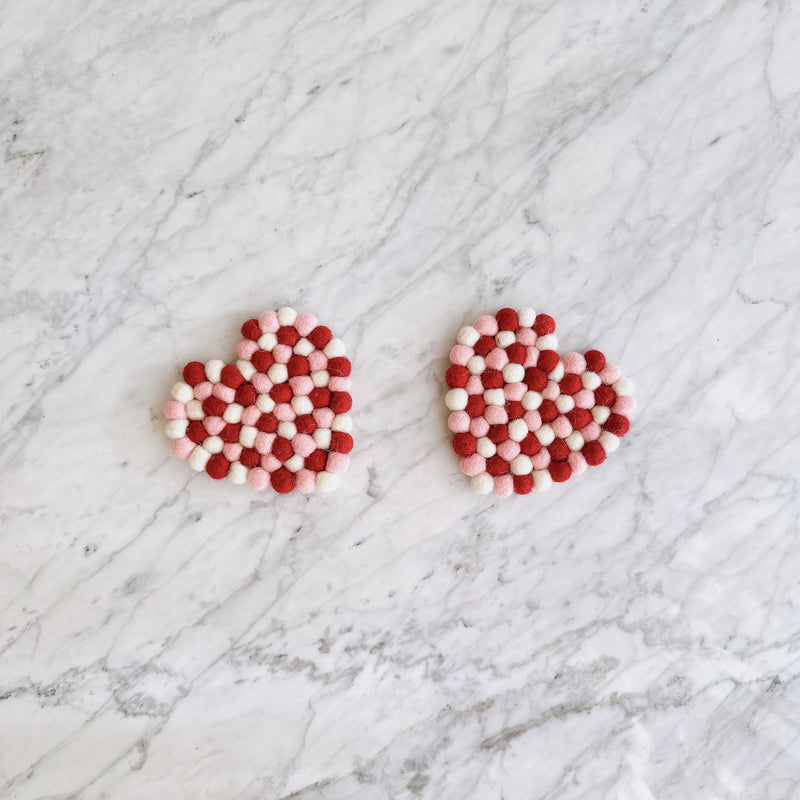 Two heart-shaped Love coasters rest on a white marble surface with gray veins. These handmade felt coasters are crafted from small, alternating red, pink, and white organic wool felt balls arranged in a grid pattern. Ethically made by Nepalese female artisans, these durable and charming coasters add a warm, romantic touch to any home decor.