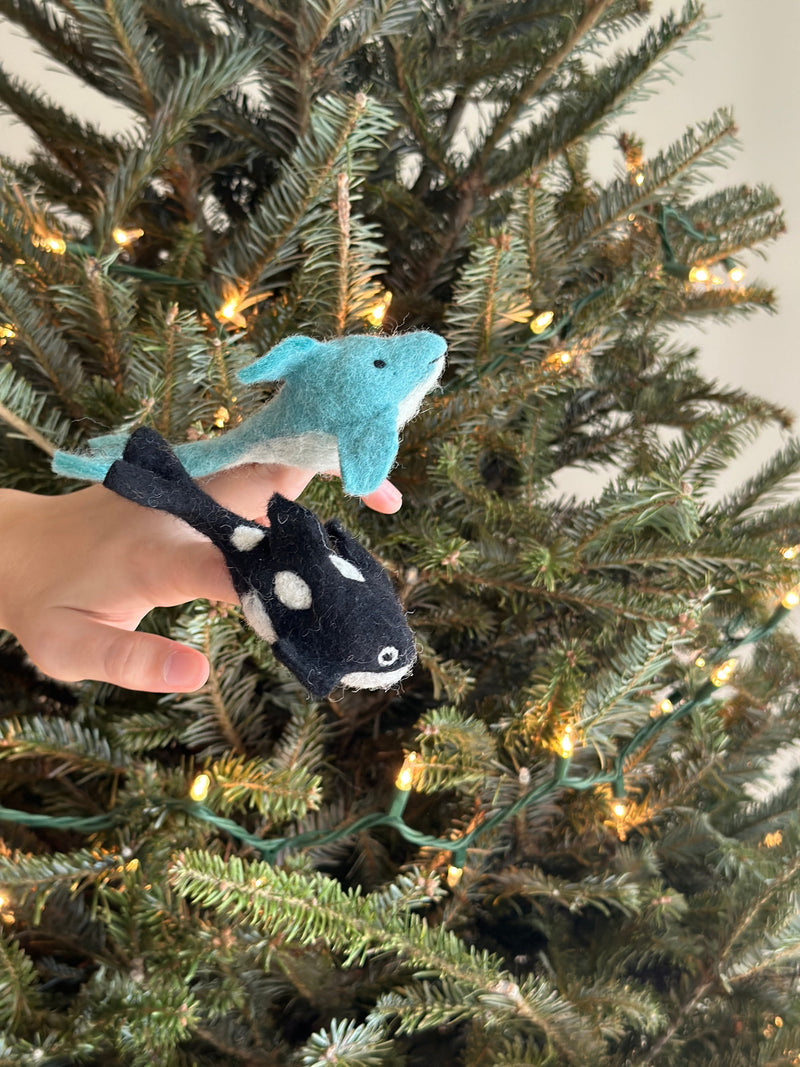 A handmade needle-felted orca finger puppet is held by a hand, prominently displayed against a blurred Christmas tree adorned with lights. This unique black and white wool felt puppet features distinct white patches around its eyes and a white belly, showcasing its detailed craftsmanship.