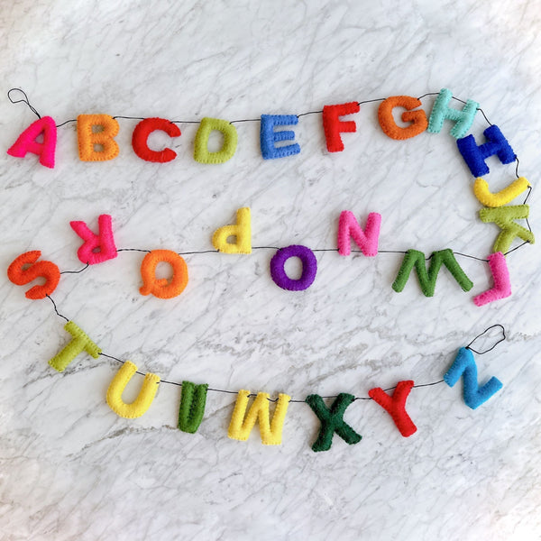A vibrant Rainbow Alphabet felt garland is cheerfully displayed on a white marble surface. This educational kids' room garland features all letters of the alphabet, from A to Z, crafted from high-quality wool felt. The letters showcase a spectrum of rainbow colors, including pink, orange, red, green, blue, yellow, and purple, strung together on a thin black cord.