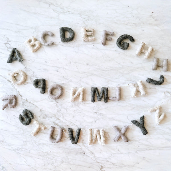 A vibrant wool felt Rainbow Alphabet Banner Kids Room Garland is displayed on a white marble surface. This garland features all 26 letters of the alphabet, each crafted in bright, eye-catching rainbow colors and strung together by a thin white string.