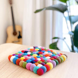 A handmade rainbow square felt ball pot trivet rests on a light wooden surface in a softly lit, homey environment, evoking a playful atmosphere. Crafted from 100% organic lamb wool, this fair-trade felt trivet features a colorful array of hand-stitched felt balls in shades of red, orange, yellow, green, blue, purple, and white.